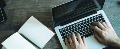 stock photo of hands typing on laptop with notebook open to the side | india paranormal author SDHegyes